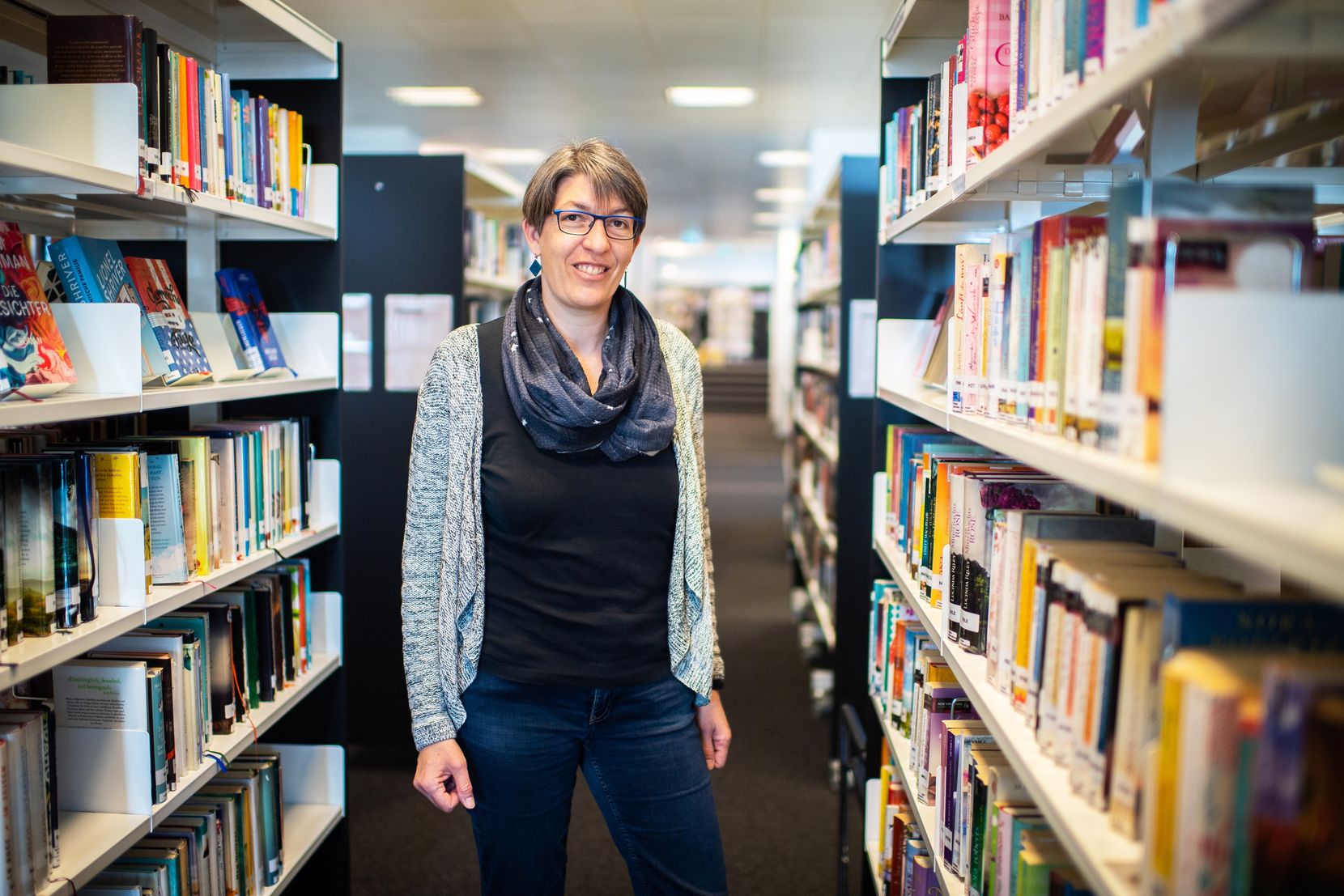 Frau mit Brille und halblangen Haaren steht in einer Bibliothek zwischen zwei Bücherregalen. 
